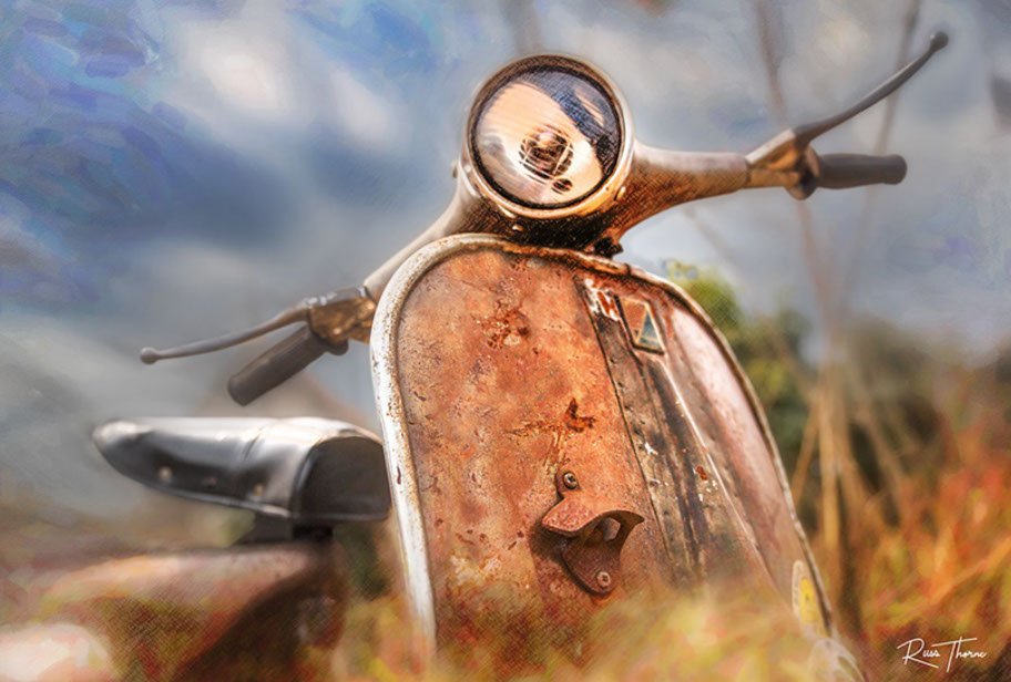 A rusting vintage Vespa scooter, partially hidden in tall weeds, reflecting years of beautiful patina.