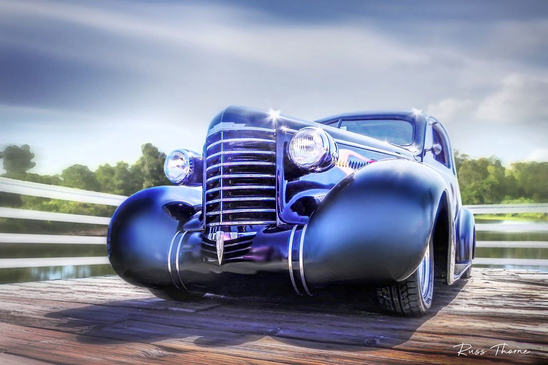 An early 1940s beautifully restored coupe parked on a pier by a tranquil lake, surrounded by lush, vibrant trees.