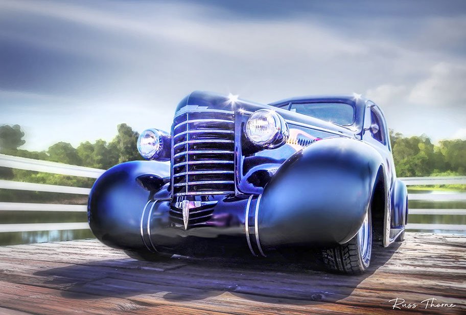 An early 1940s beautifully restored coupe parked on a pier by a tranquil lake, surrounded by lush, vibrant trees.