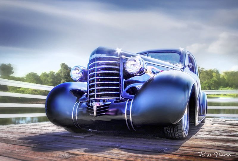 An early 1940s beautifully restored coupe parked on a pier by a tranquil lake, surrounded by lush, vibrant trees.