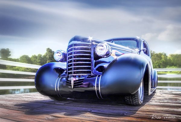 An early 1940s beautifully restored coupe parked on a pier by a tranquil lake, surrounded by lush, vibrant trees.